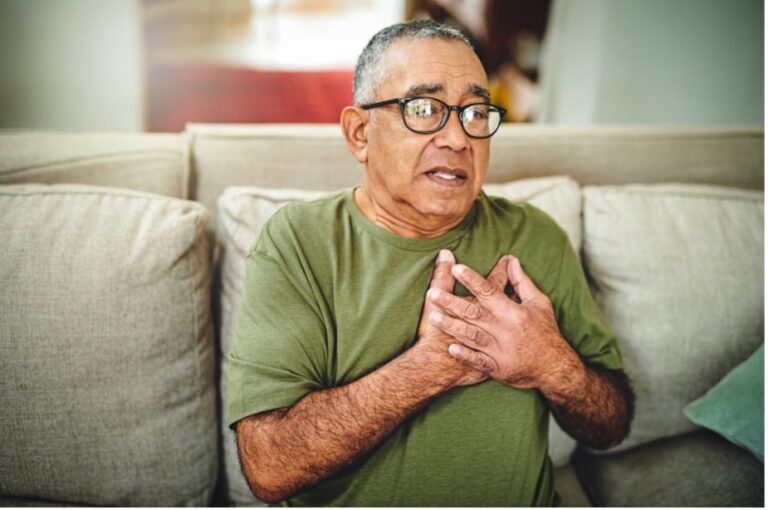 A senior man sits on the sofa, holding his chest in pain caused by consuming a high level of fructose